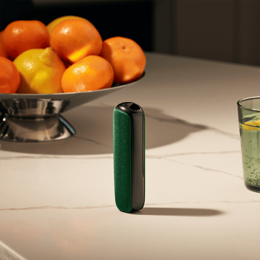 Ploom AURA uređaj sa Amazon Green zadnjom maskom na stolu pored korpe sa voćem